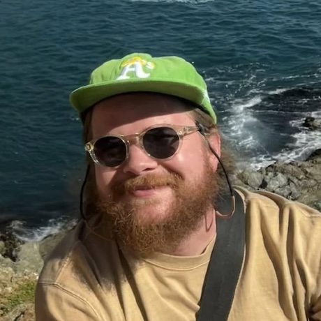 Gavin in sunglasses and a green ball cap, ocean behind him